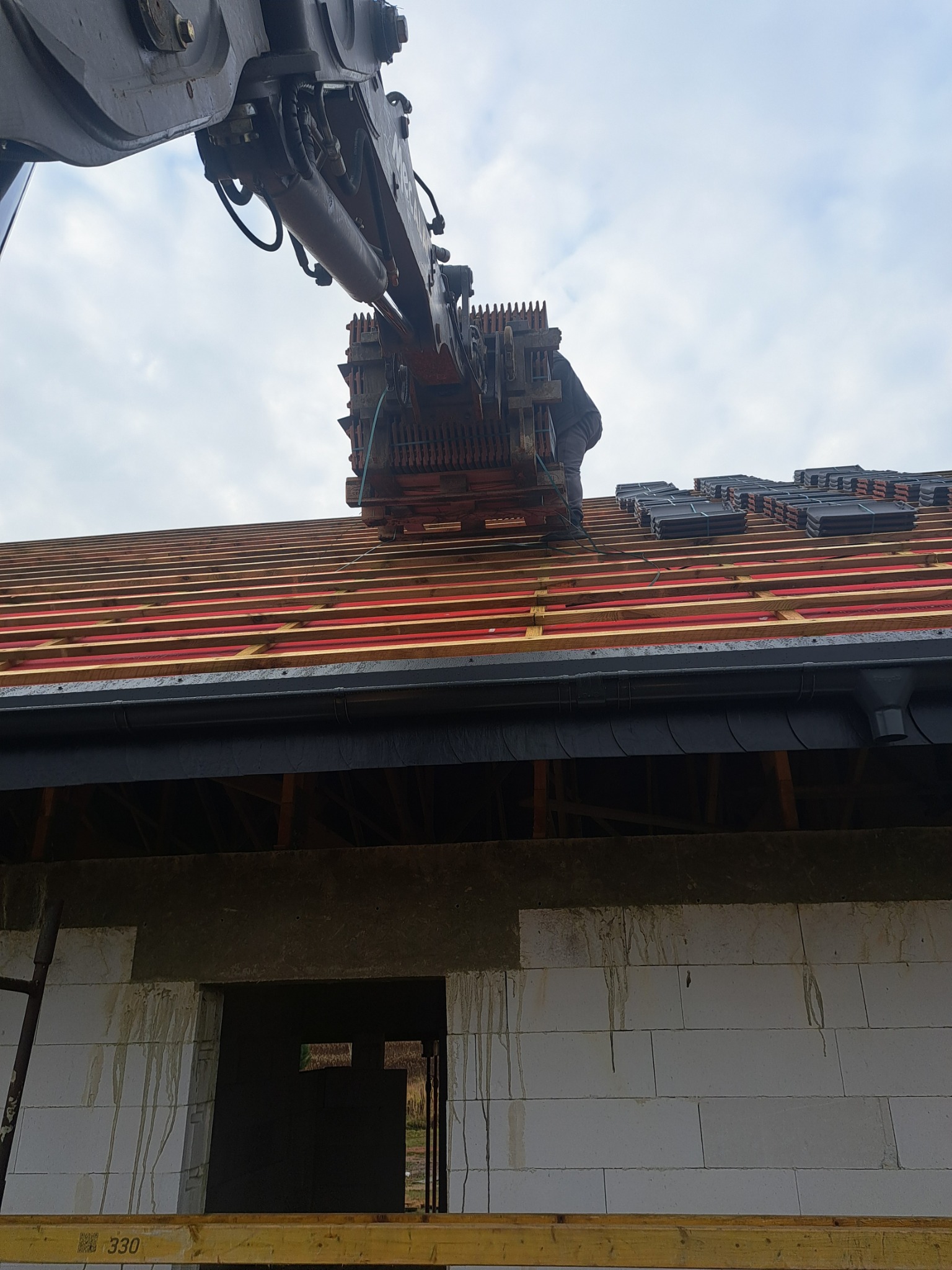 Żuraw podnosi paletę z dachówkami na drewnianą konstrukcję dachu, widoczny fragment ściany budynku z białych bloczków i niezakończonym ociepleniem.