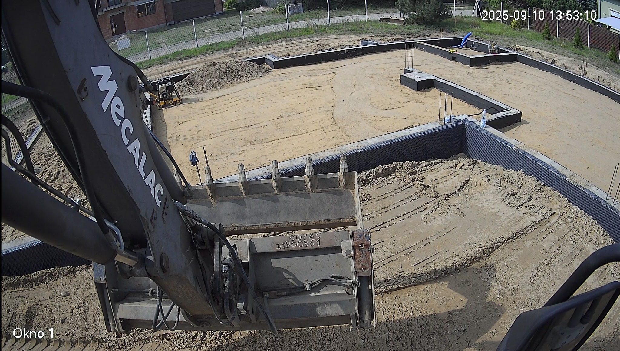 Praca ziemna z użyciem koparki Mecalac. Wykop pod fundamenty z widoczną hydroizolacją, zagęszczarka w tle. Ujęcie z kabiny operatora.