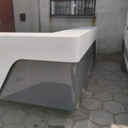 Nowoczesna, dwukolorowa lada recepcyjna z białym blatem i szarym korpusem, ustawiona na zewnątrz budynku na kostce brukowej, widoczna z boku pod kątem.