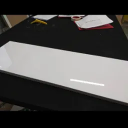 Biały, lakierowany front meblowy leży na czarnym blacie roboczym, widoczne narzędzia i dokumenty w tle.