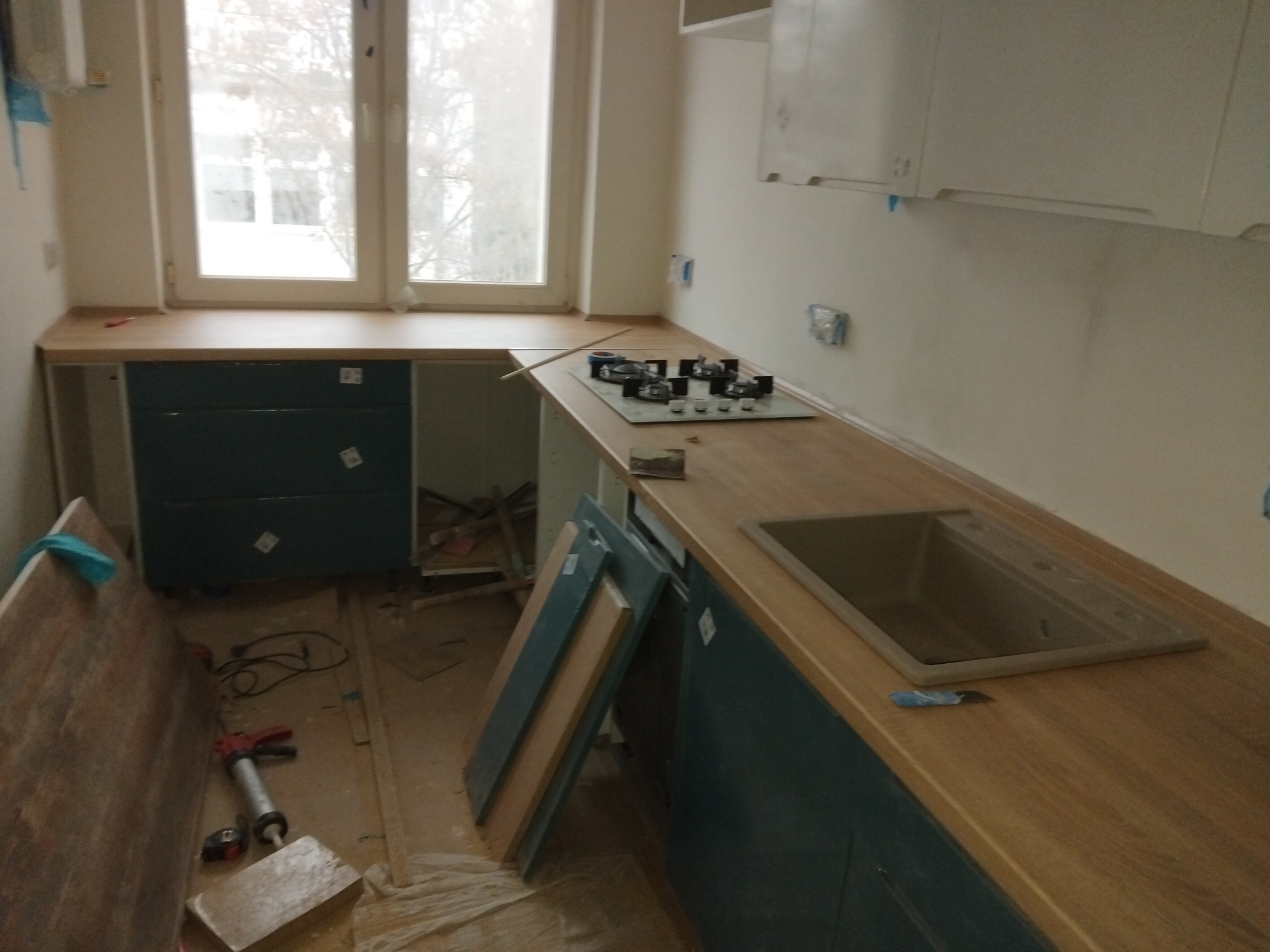Montaż kuchni: widoczne niedokończone szafki dolne w kolorze ciemnego turkusu, blat z jasnego drewna, płyta grzewcza i zlew w trakcie instalacji, narzędzia i materiały budowlane na podłodze.