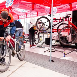 Mobilny Serwis Rowerowy Dr Albin - Mobilny serwis rowerowy: mężczyzna w okularach i kurtce naprawia rower górski na chodniku, pod czerwonym namiotem z logo serwisu, w tle widoczne inne rowery i pracownicy.