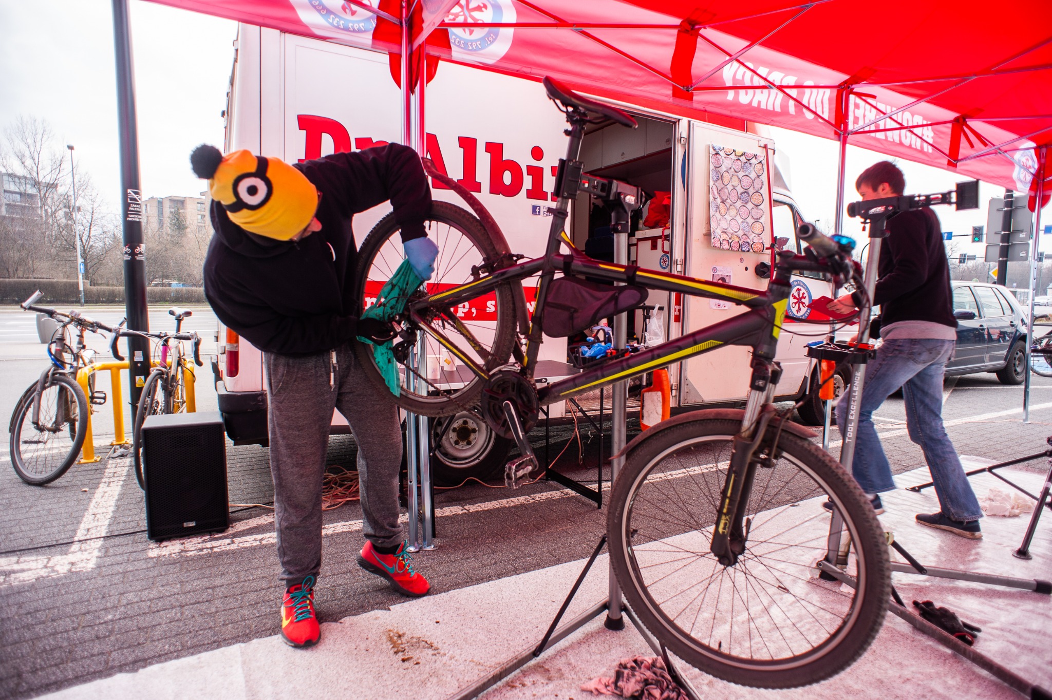 Mobilny serwis rowerowy: mechanik w czapce Minionka czyści rower na stojaku, w tle bus serwisowy z logo i drugi mechanik.