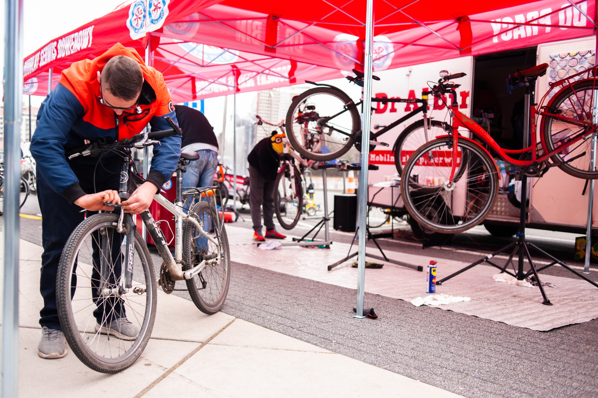 Mobilny serwis rowerowy: mężczyzna w okularach i kurtce naprawia rower górski na chodniku, pod czerwonym namiotem z logo serwisu, w tle widoczne inne rowery i pracownicy.
