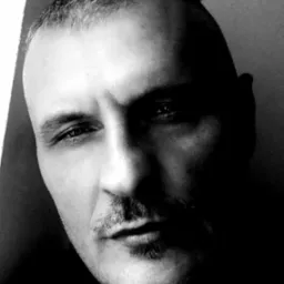 Czarno-biały portret mężczyzny z wąsami, oświetlonego bocznym światłem, tworzącym silne kontrasty i cienie na twarzy.