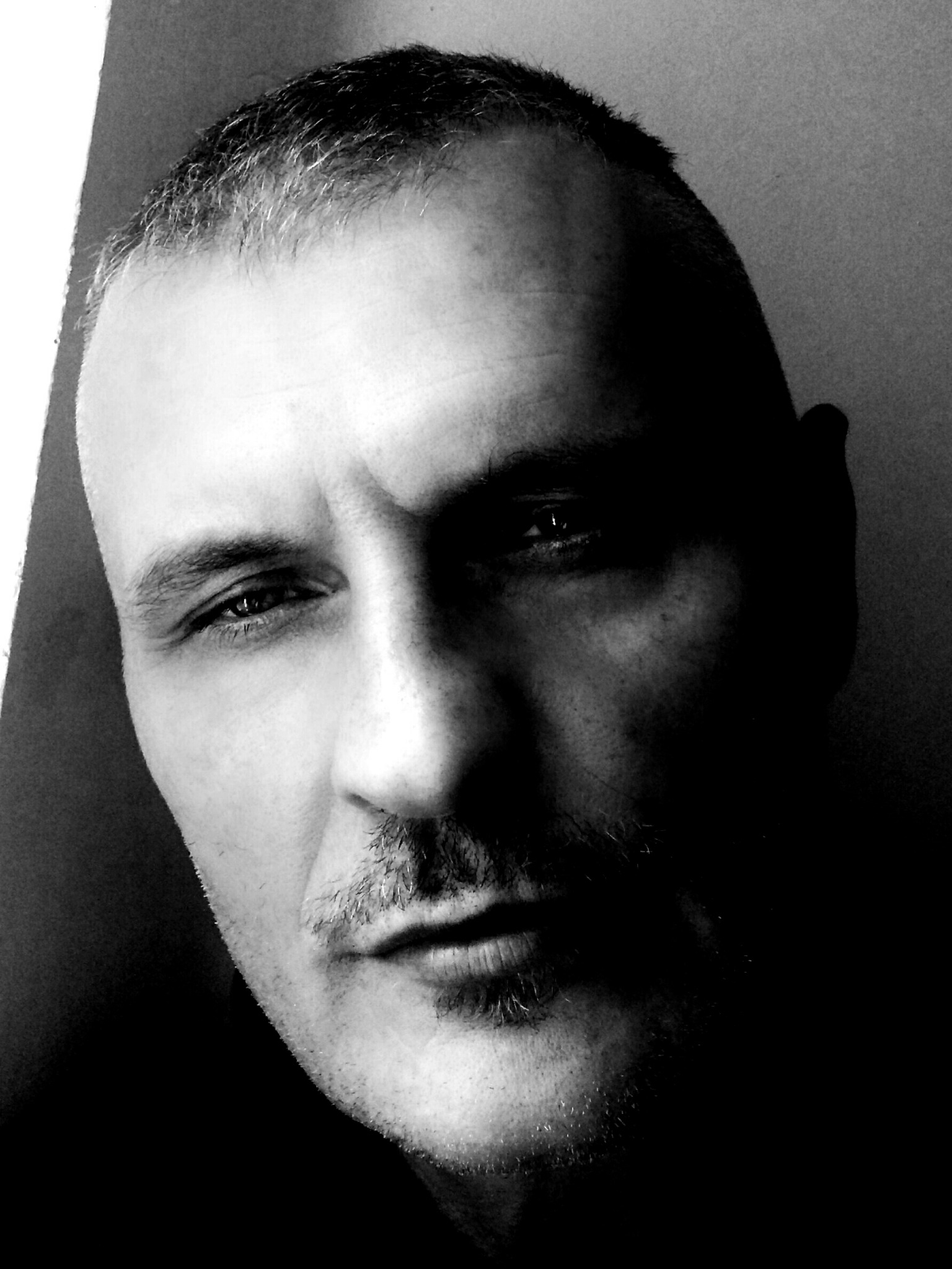 Czarno-biały portret mężczyzny z wąsami, oświetlonego bocznym światłem, tworzącym silne kontrasty i cienie na twarzy.