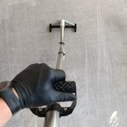 Mobifresh - Pranie jasnoszarego dywanu metodą ekstrakcyjną, widoczna ręka w czarnej rękawicy trzymająca uchwyt odkurzacza piorącego, fragment dywanu już wyczyszczony.