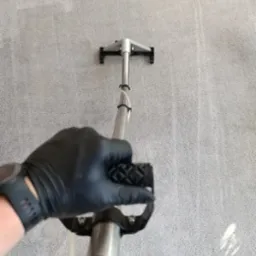 Pranie jasnoszarego dywanu metodą ekstrakcyjną, widoczna ręka w czarnej rękawicy trzymająca uchwyt odkurzacza piorącego, fragment dywanu już wyczyszczony.