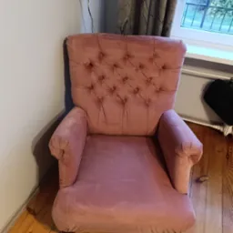 Różowy, pluszowy fotel typu uszak z guzikami na oparciu, stojący na drewnianej podłodze, z widocznym fragmentem dywanu i okna w tle.