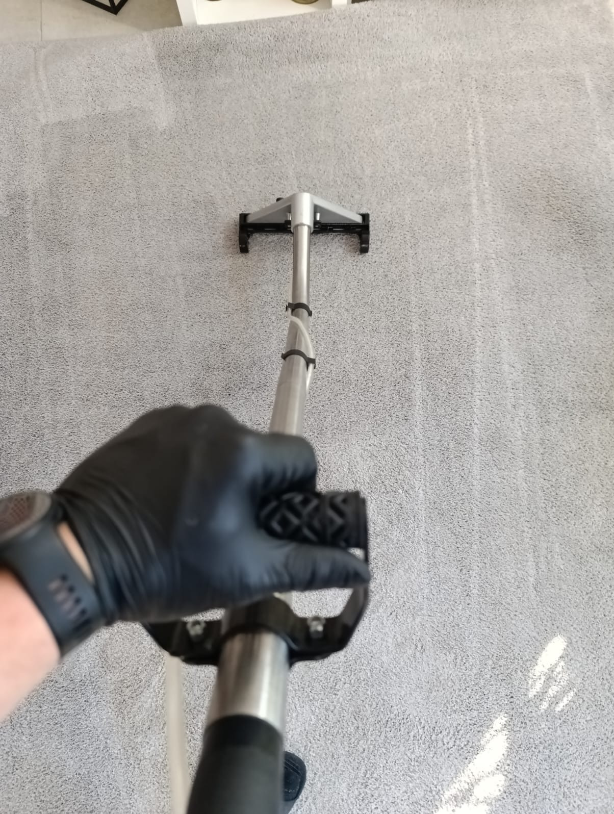 Pranie jasnoszarego dywanu metodą ekstrakcyjną, widoczna ręka w czarnej rękawicy trzymająca uchwyt odkurzacza piorącego, fragment dywanu już wyczyszczony.