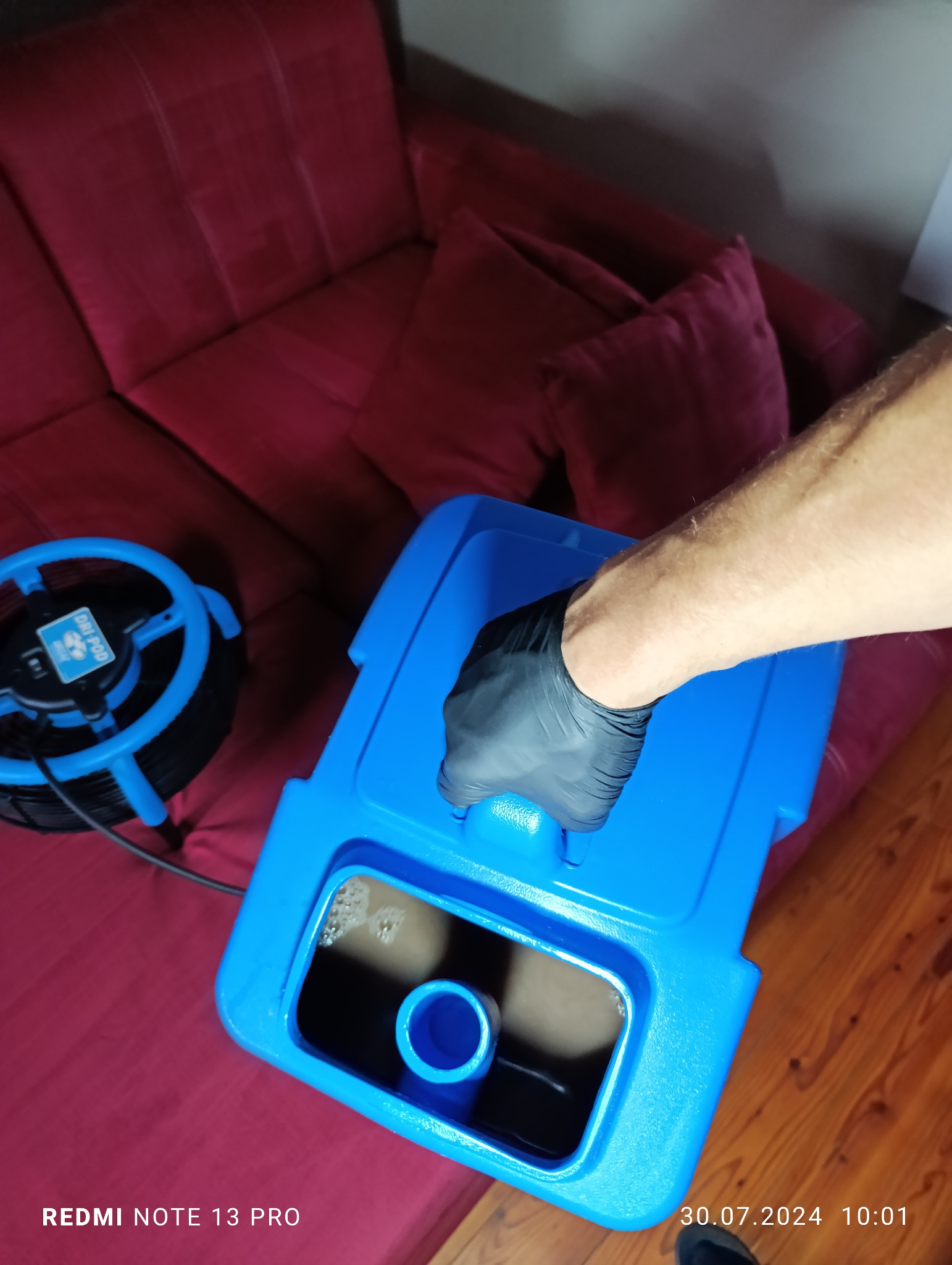 Pracownik w czarnej rękawicy trzyma niebieski pojemnik z brudną wodą po czyszczeniu, obok widoczny sprzęt czyszczący i fragment czerwonej sofy.