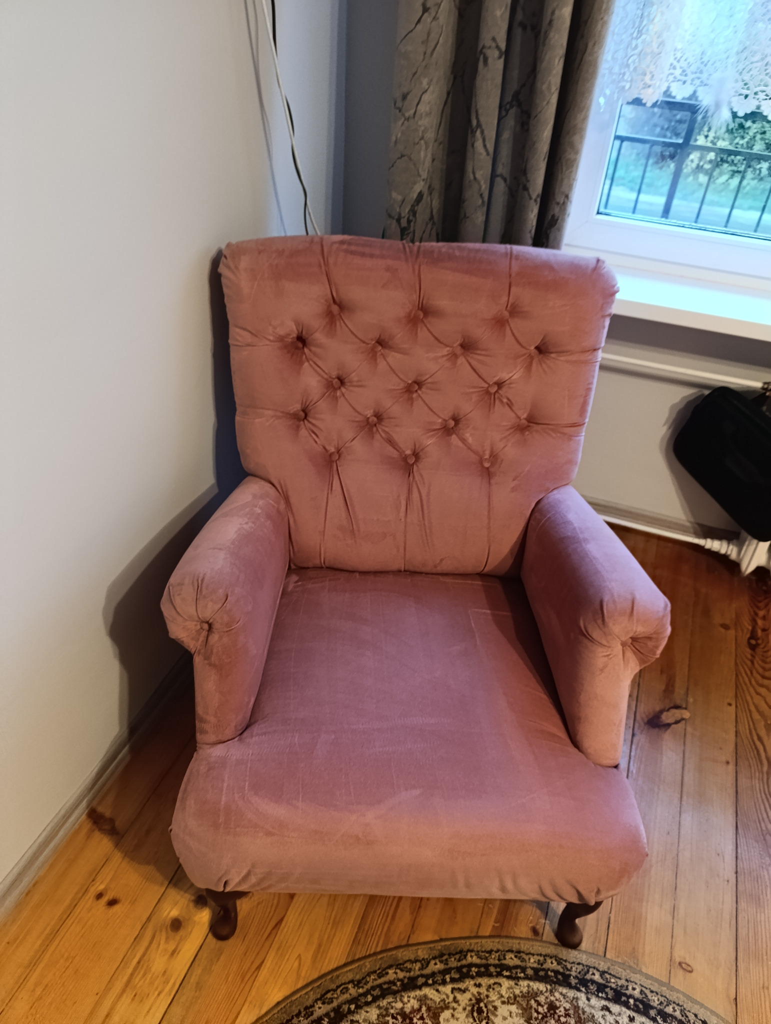 Różowy, pluszowy fotel typu uszak z guzikami na oparciu, stojący na drewnianej podłodze, z widocznym fragmentem dywanu i okna w tle.