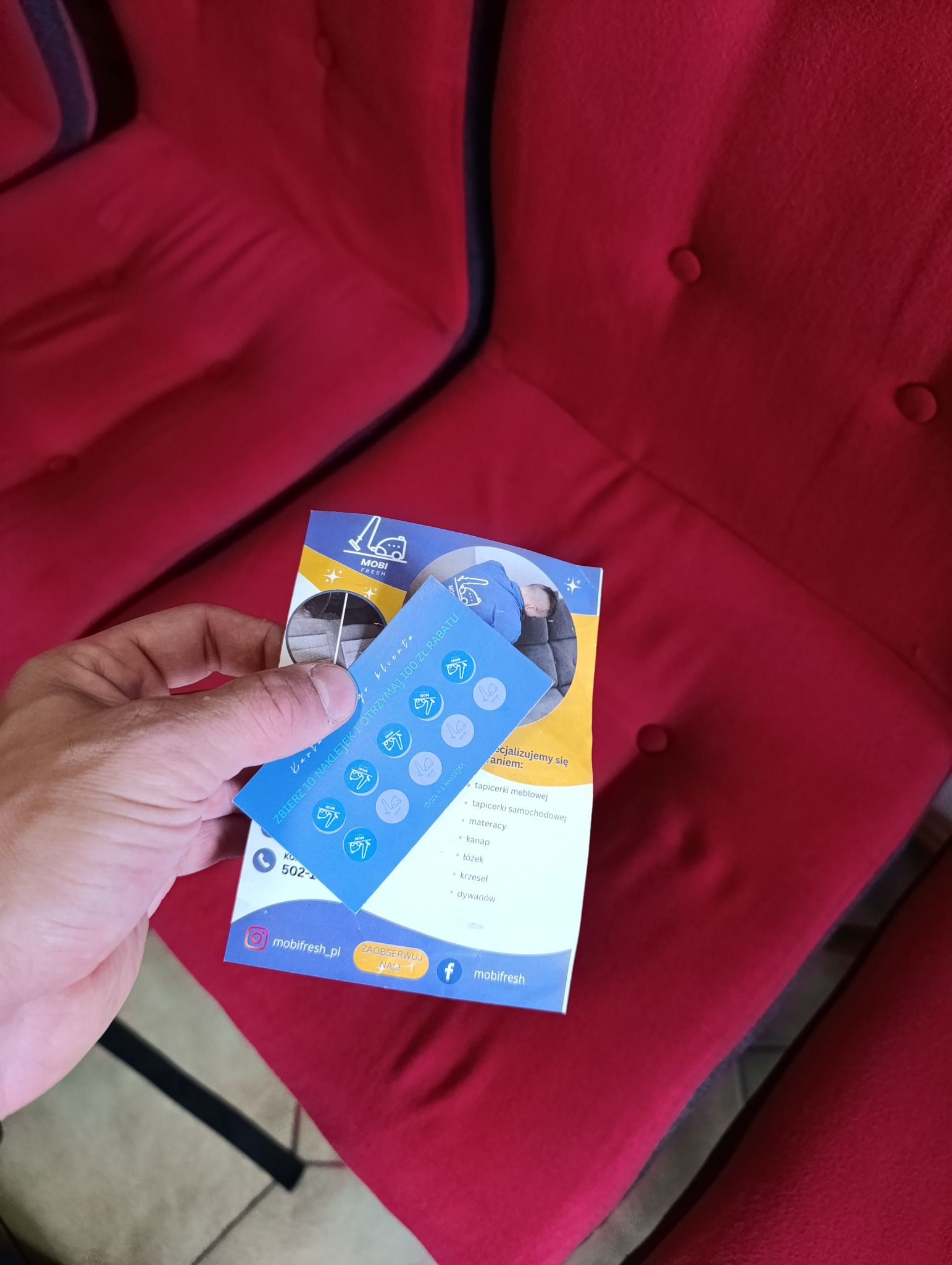 Czerwona sofa z guzikami w tle, na pierwszym planie ulotka reklamowa firmy oferującej usługi czyszczenia tapicerki meblowej, samochodowej, materacy, kanap, łóżek, krzeseł i dywanów.