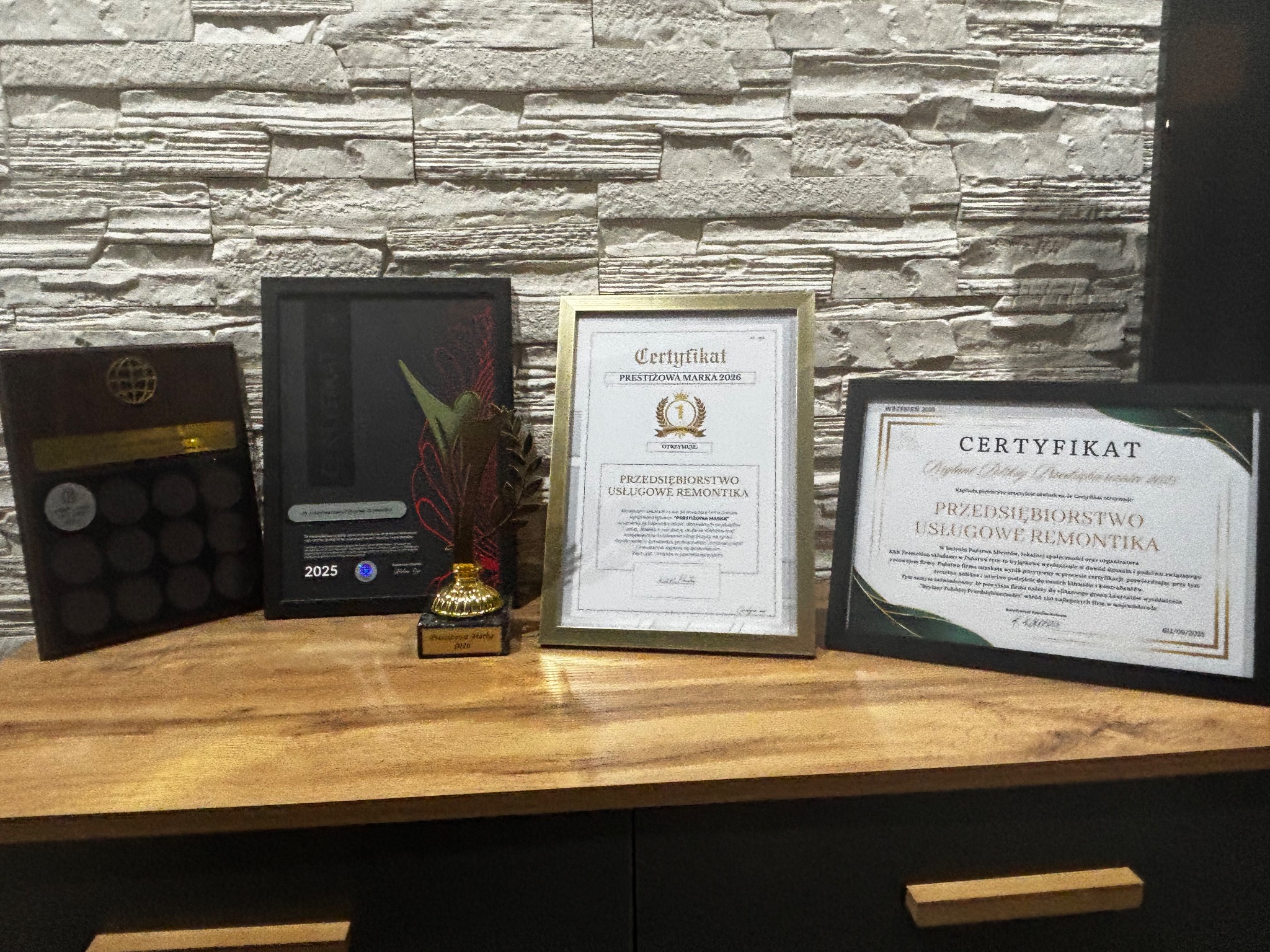 Certyfikaty i nagrody firmy remontowej na drewnianym blacie, na tle ściany z dekoracyjnej cegły. Trofea z lat 2025 i 2026.
