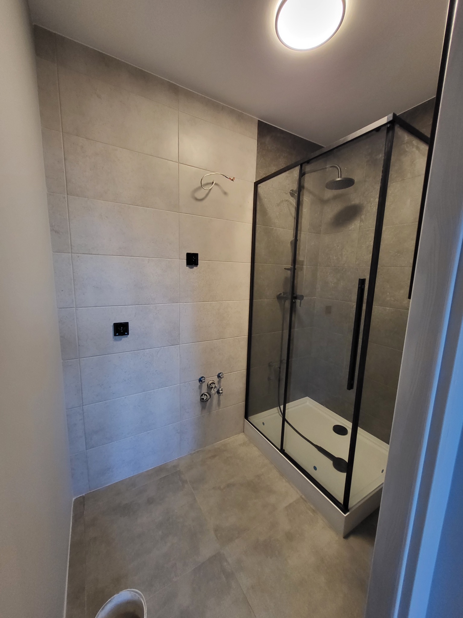 Wykończona łazienka z kabiną prysznicową typu walk-in z czarnymi profilami, szare płytki na ścianach i podłodze, widoczne przyłącza wodne i instalacja elektryczna.