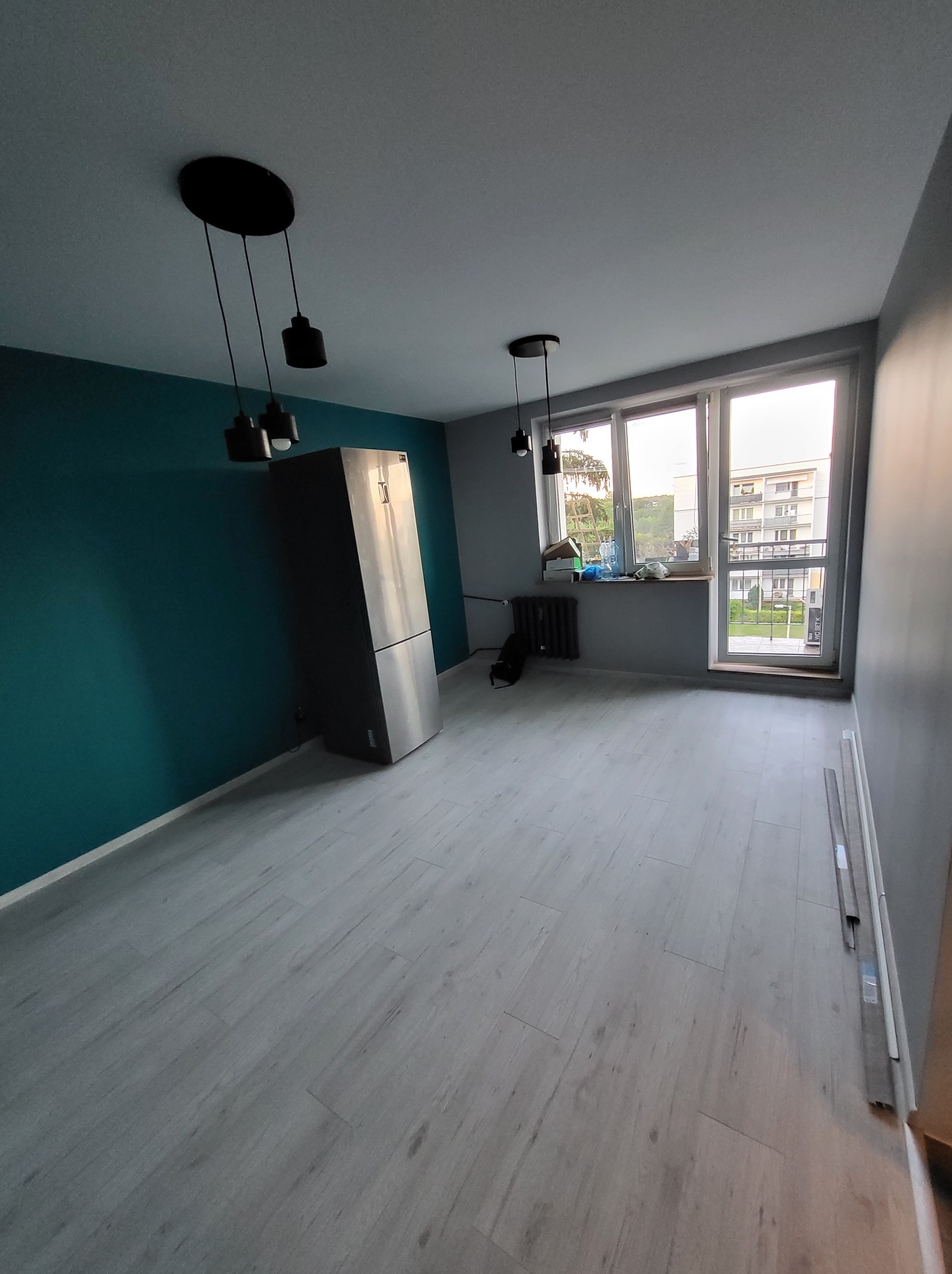 Pomalowane na szaro i turkusowo wnętrze pokoju z panelami podłogowymi, z oknem i grzejnikiem. Widoczne lampy sufitowe i lodówka. Świeżo po malowaniu.