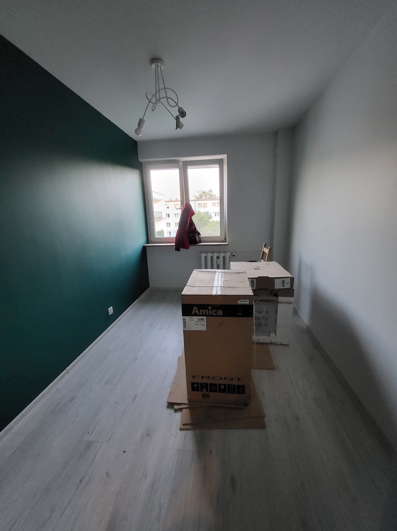 Pomalowany na butelkową zieleń fragment ściany w pokoju z podłogą w panelach. W tle okno i kartony z nowym sprzętem AGD. Nowe mieszkanie po remoncie.