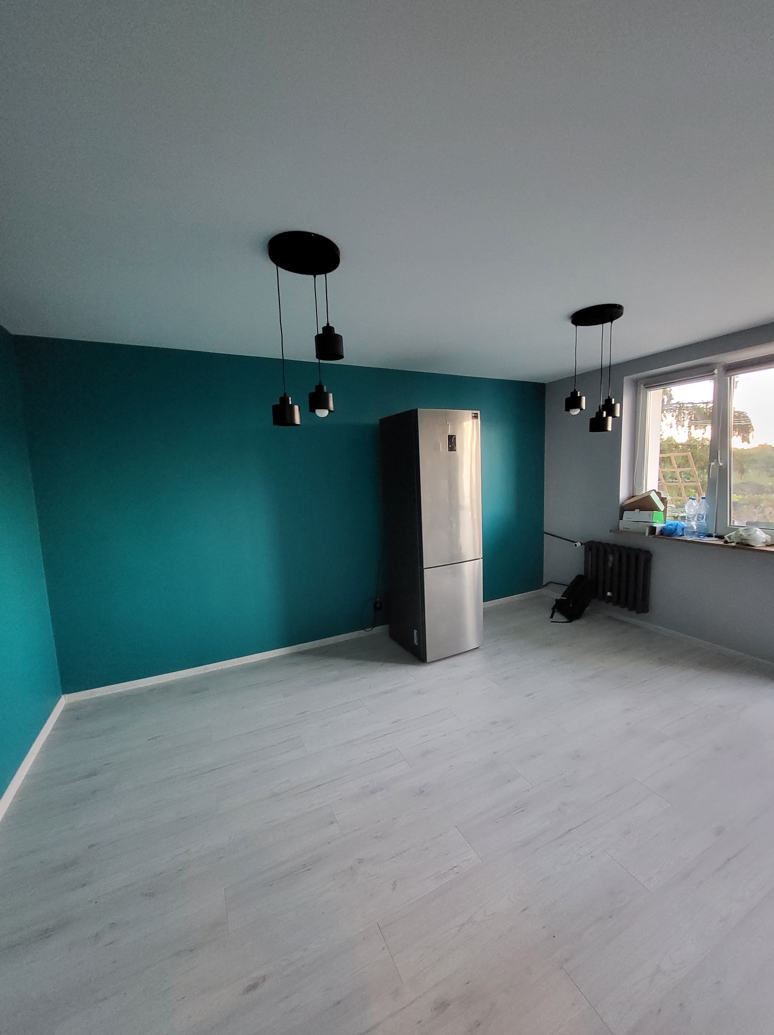 Pomalowane na turkusowo ściany w pokoju z szarą podłogą i oknem. Widoczne czarne lampy sufitowe i srebrna lodówka. Świeżo po malowaniu, czysto i schludnie.