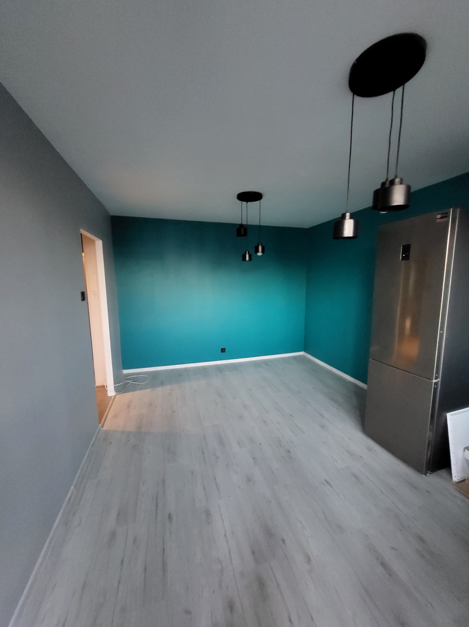 Pomalowane na turkusowo ściany w pustym pokoju z szarą podłogą i trzema nowoczesnymi lampami wiszącymi. Widoczna lodówka i fragment ściany pomalowanej na szaro.