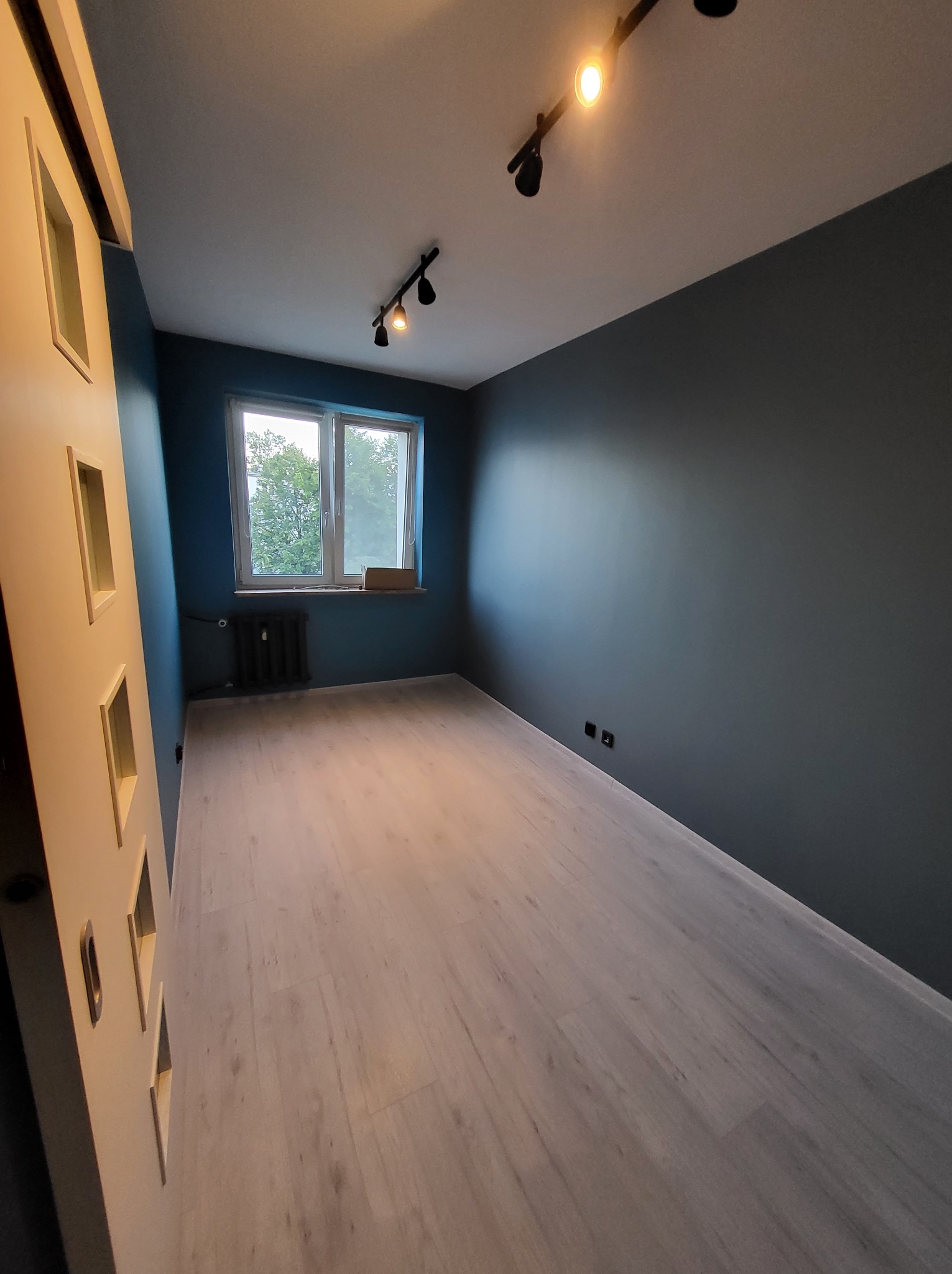 Pomalowane na ciemny kolor wnętrze pokoju z jasną podłogą i oknem. Widoczne oświetlenie sufitowe i dekoracyjne drzwi przesuwne z wycięciami.