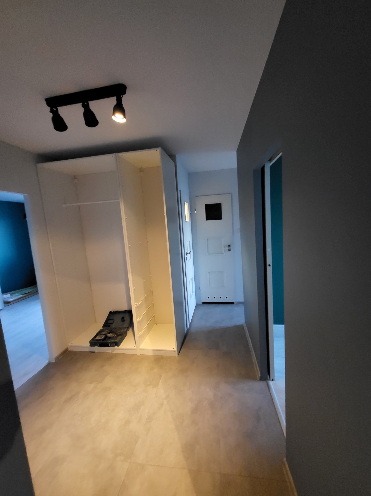 Wnętrze korytarza z zamontowaną białą szafą wnękową, widoczne narzędzia, szara podłoga i ściany. Oświetlenie sufitowe punktowe. Drzwi w tle.