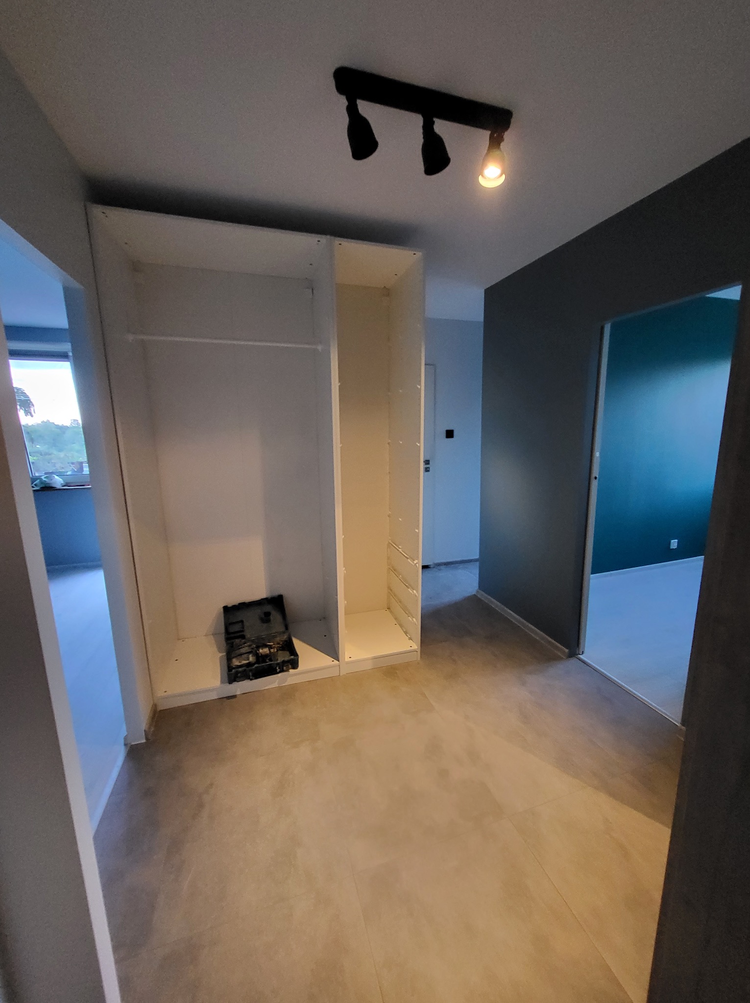 Wnętrze w trakcie wykańczania: biała szafa wnękowa z narzędziami, szare ściany i podłoga z płytek. Widok na sąsiednie pomieszczenia.