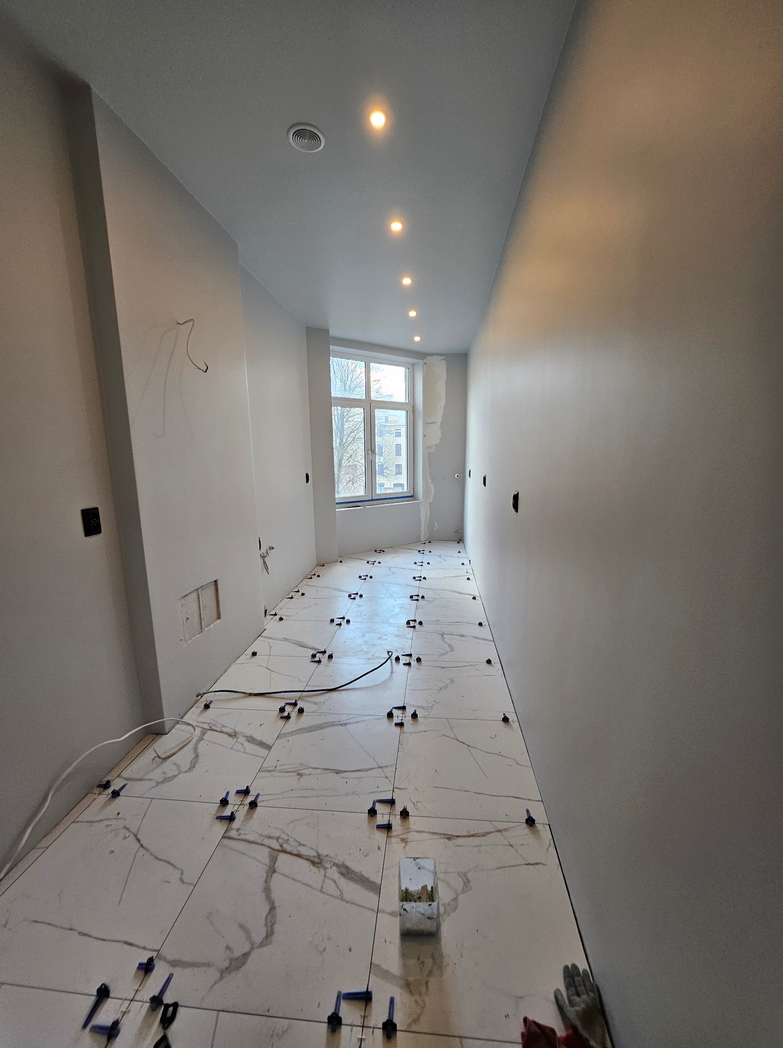 Układanie płytek podłogowych w Lubieniu. Białe płytki imitujące marmur z niebieskimi krzyżykami dystansowymi. Świeżo wykończone ściany i sufit z oświetleniem.