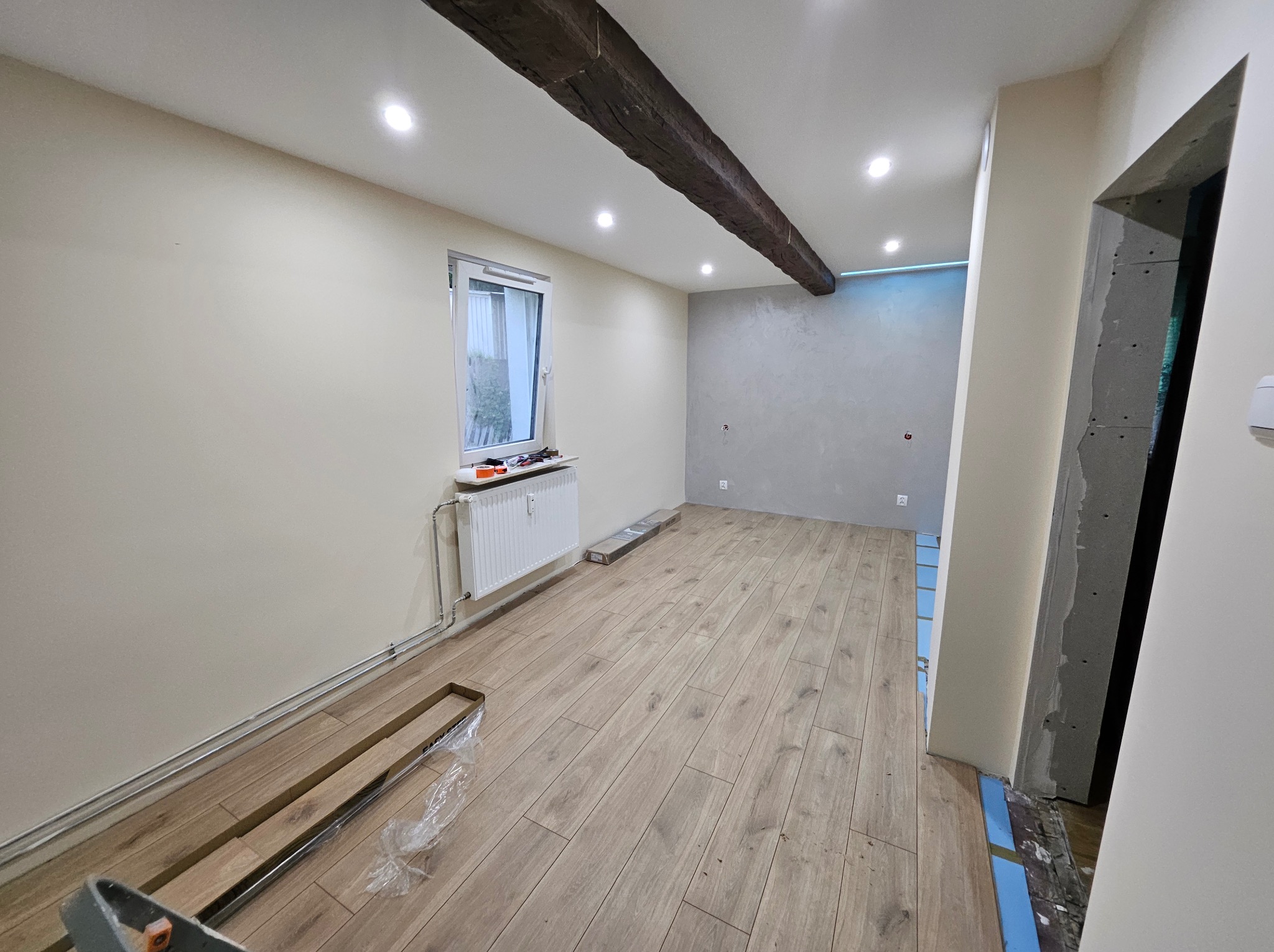 Jasne panele podłogowe ułożone w pokoju z beżowymi ścianami i drewnianą belką stropową. Widoczne narzędzia i materiały budowlane. Częściowo wykończone wnętrze.