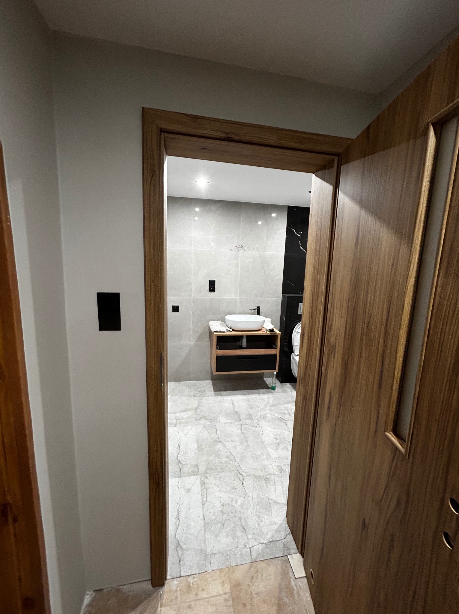 Nowoczesna łazienka z marmurową podłogą i szarą glazurą, widoczna przez drewniane drzwi. Umywalka nablatowa na szafce z ciemnego drewna. Minimalistyczny design.