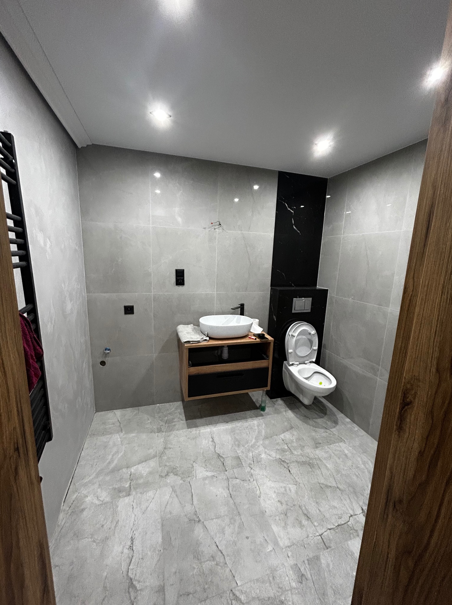 Nowoczesna łazienka z szarymi płytkami imitującymi marmur, wiszącą szafką z umywalką i toaletą. Czarny panel dekoracyjny dodaje elegancji. Widok zza drewnianej framugi.