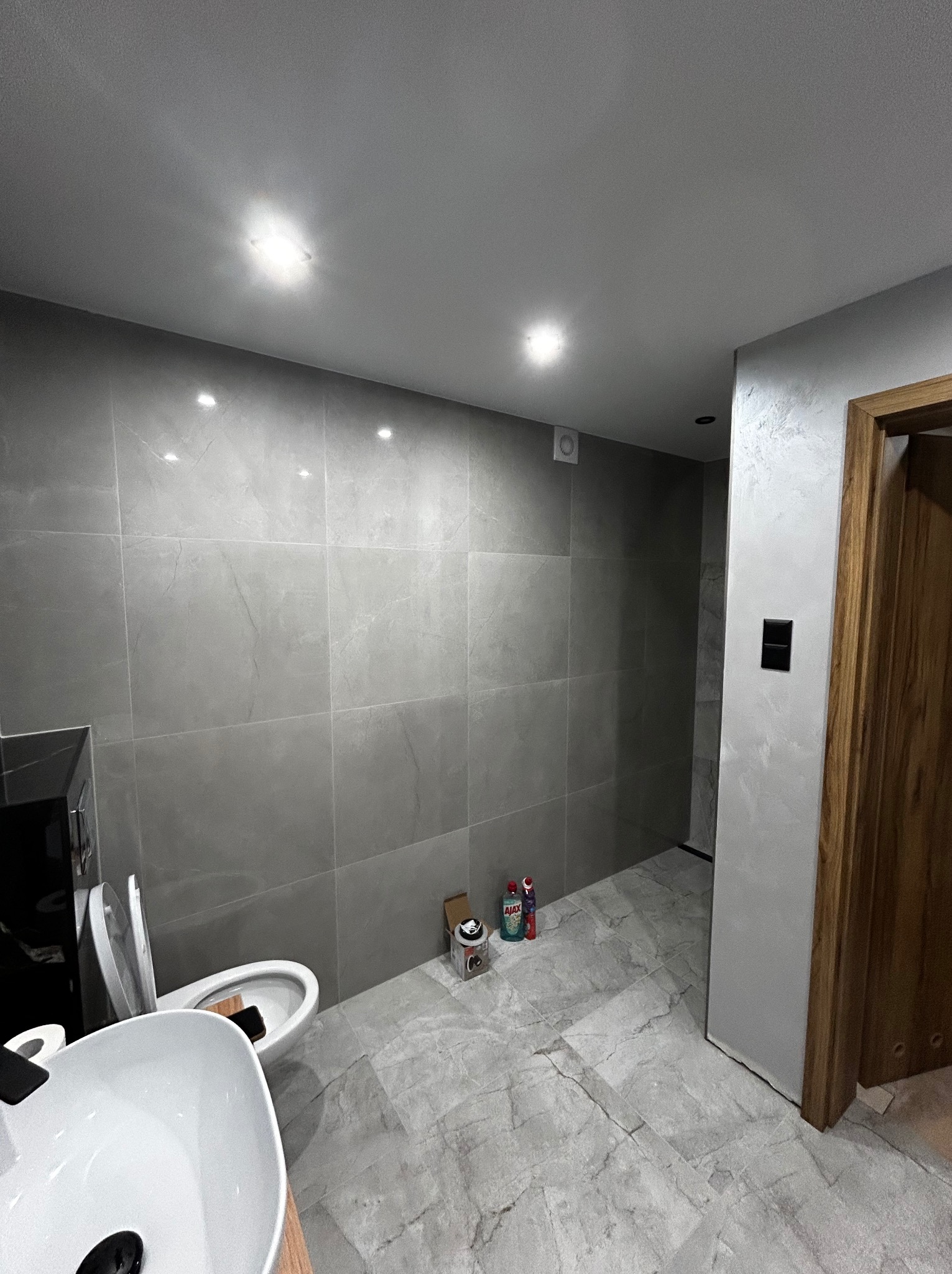 Nowoczesna łazienka z szarymi płytkami imitującymi marmur na ścianach i podłodze. Umywalka nablatowa, toaleta i drewniana rama drzwi. Oświetlenie punktowe.