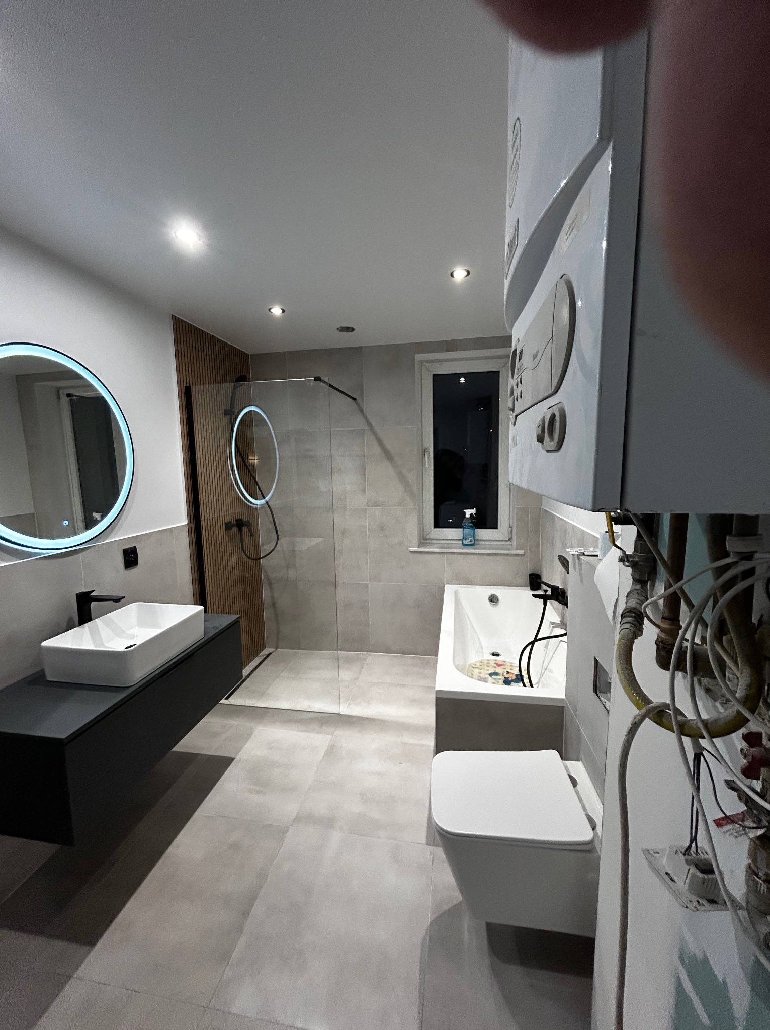 Nowoczesna łazienka z podświetlanym lustrem, kabiną prysznicową typu walk-in i wiszącą szafką z umywalką. Widoczny bojler i okno z butelką płynu. Szare płytki i drewniane lamele.