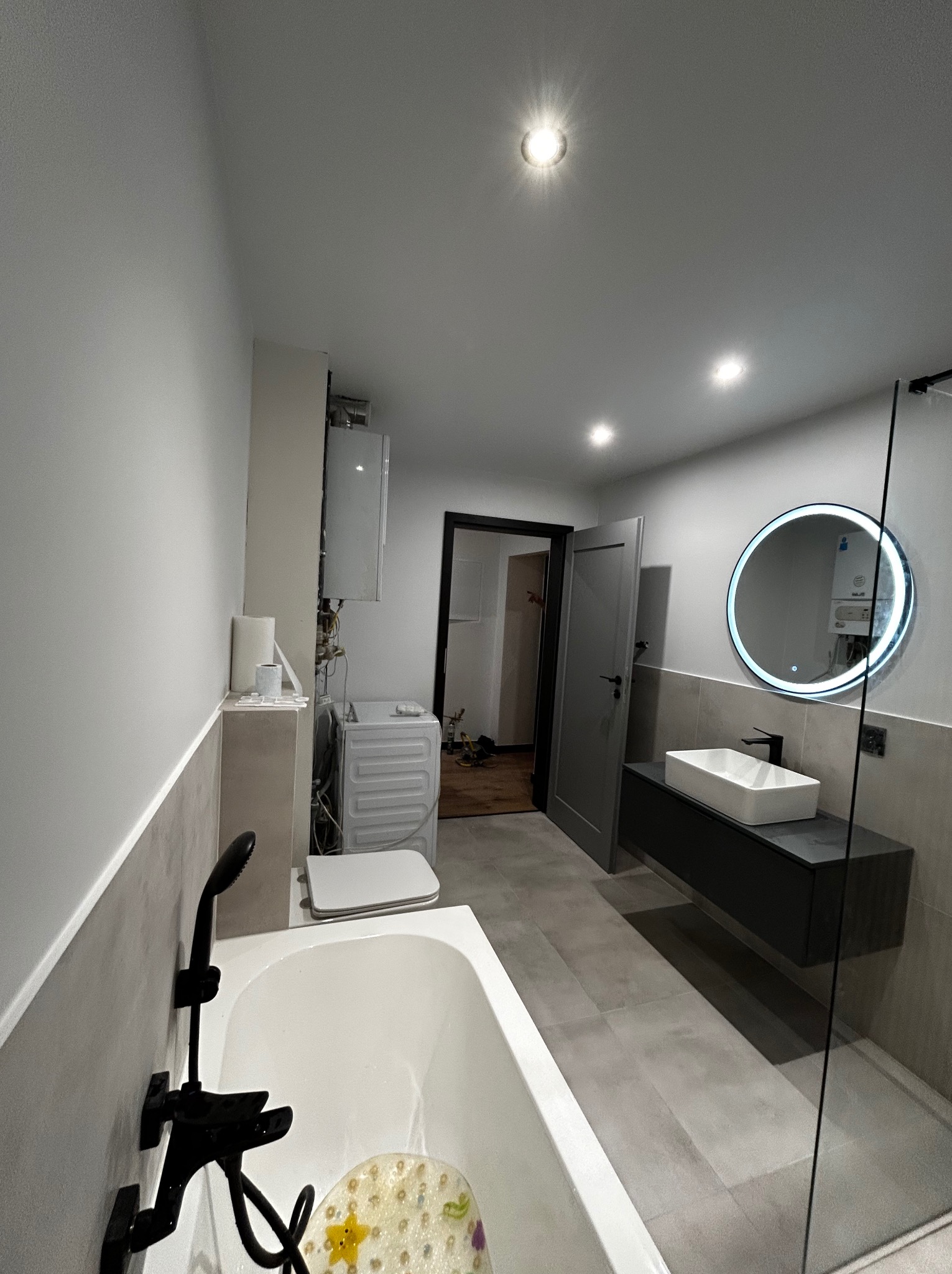 Nowoczesna łazienka po remoncie: szare płytki, biała wanna z czarną armaturą, podświetlane lustro, umywalka na szafce. Widoczna pralka i bojler.