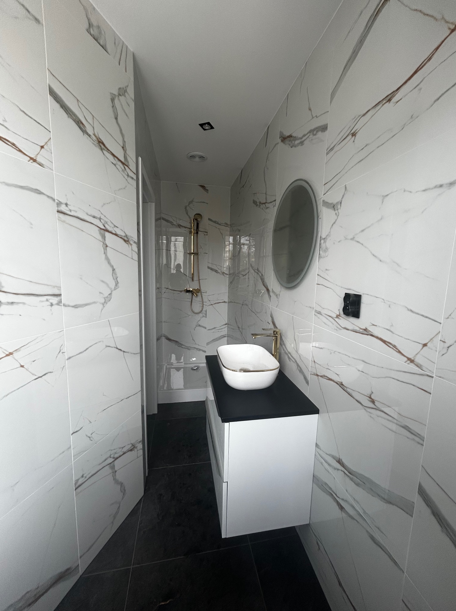 Nowoczesna łazienka z białymi płytkami imitującymi marmur, czarną podłogą i blatem. Umywalka nablatowa w kolorze białym, złota armatura i okrągłe lustro.