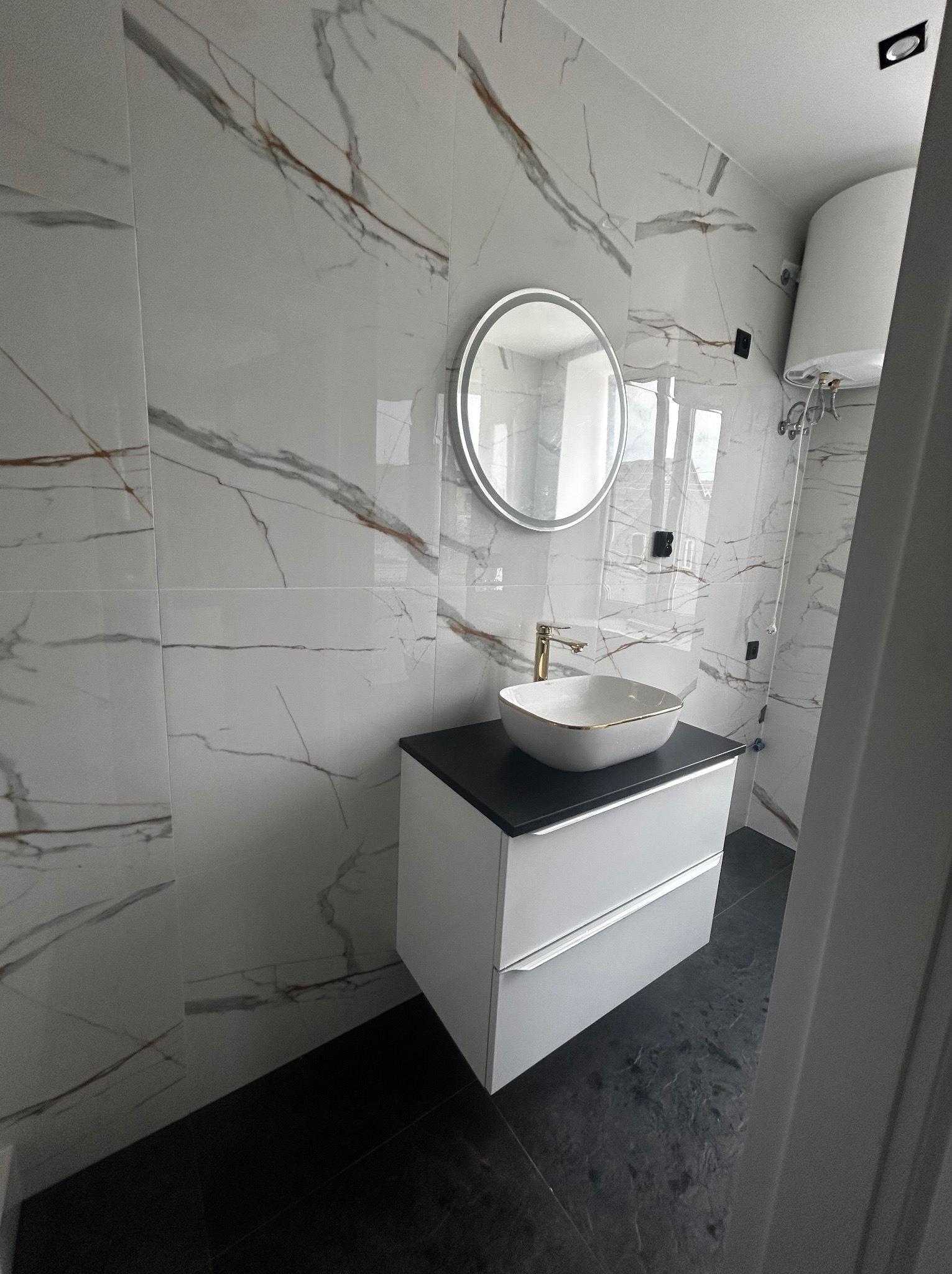 Nowoczesna łazienka z umywalką nablatową, okrągłym lustrem i białymi płytkami imitującymi marmur z brązowymi żyłami. Czarna podłoga i biała szafka pod umywalką.