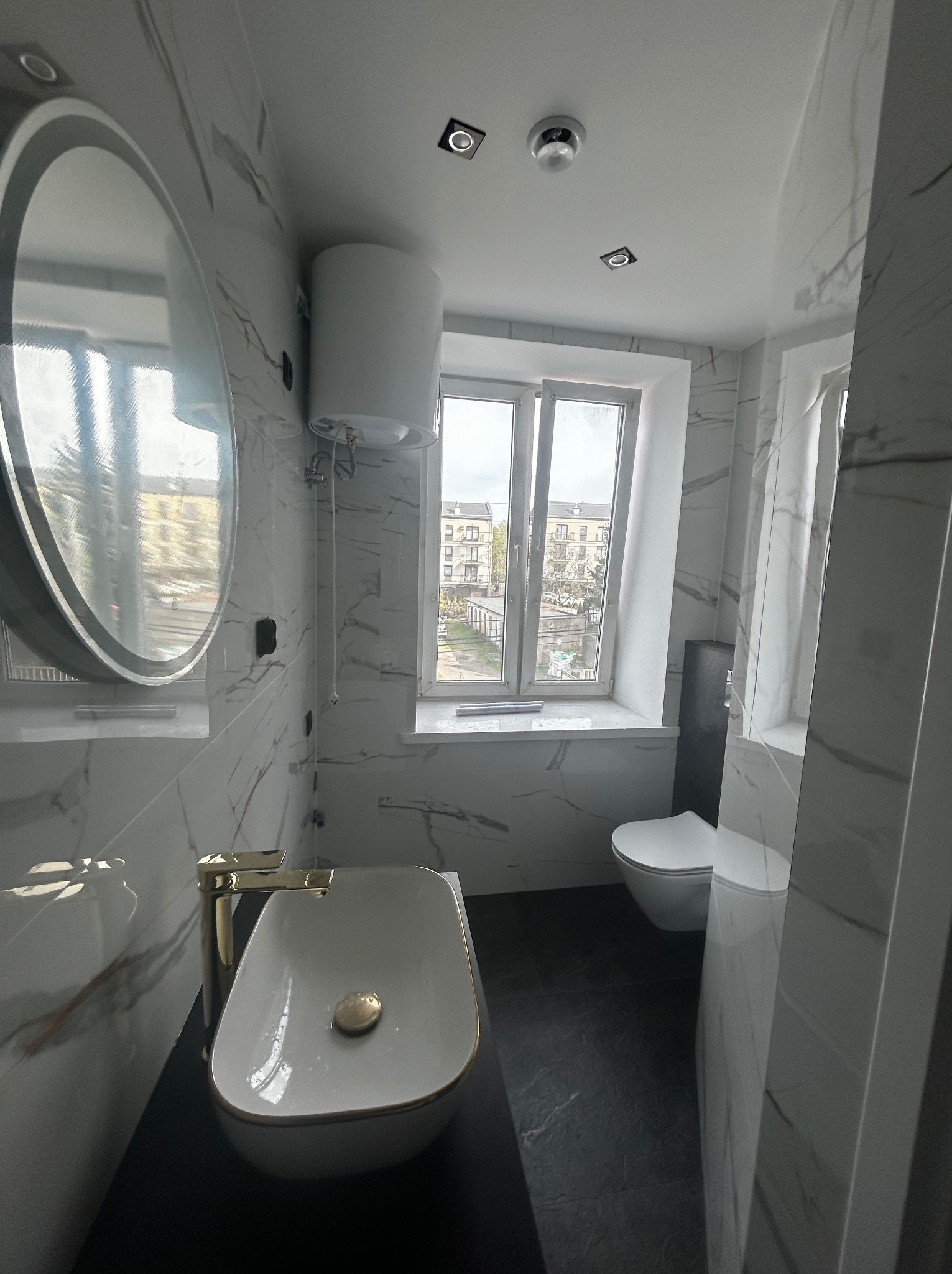 Wykończona łazienka z marmurowymi płytkami, owalną umywalką z baterią w kolorze złota i dużym lustrem. Widoczne okno z widokiem na zewnątrz.