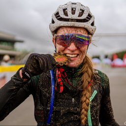 Kacper Loch Photography - Uśmiechnięta, brudna od błota kobieta w kasku i okularach przeciwsłonecznych, trzymająca w dłoni medal z numerem 60 na tle mety wyścigu.
