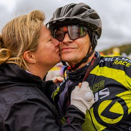 Kacper Loch Photography - Kobieta całuje w policzek mężczyznę w kasku rowerowym i okularach przeciwsłonecznych, oboje ubrani w odzież sportową Kellys Race, widoczne zabrudzenia błotem.