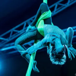 Akrobatka w kostiumie, zwisająca głową w dół na zielonej chuście, oświetlona niebieskim światłem reflektorów na tle konstrukcji scenicznej.