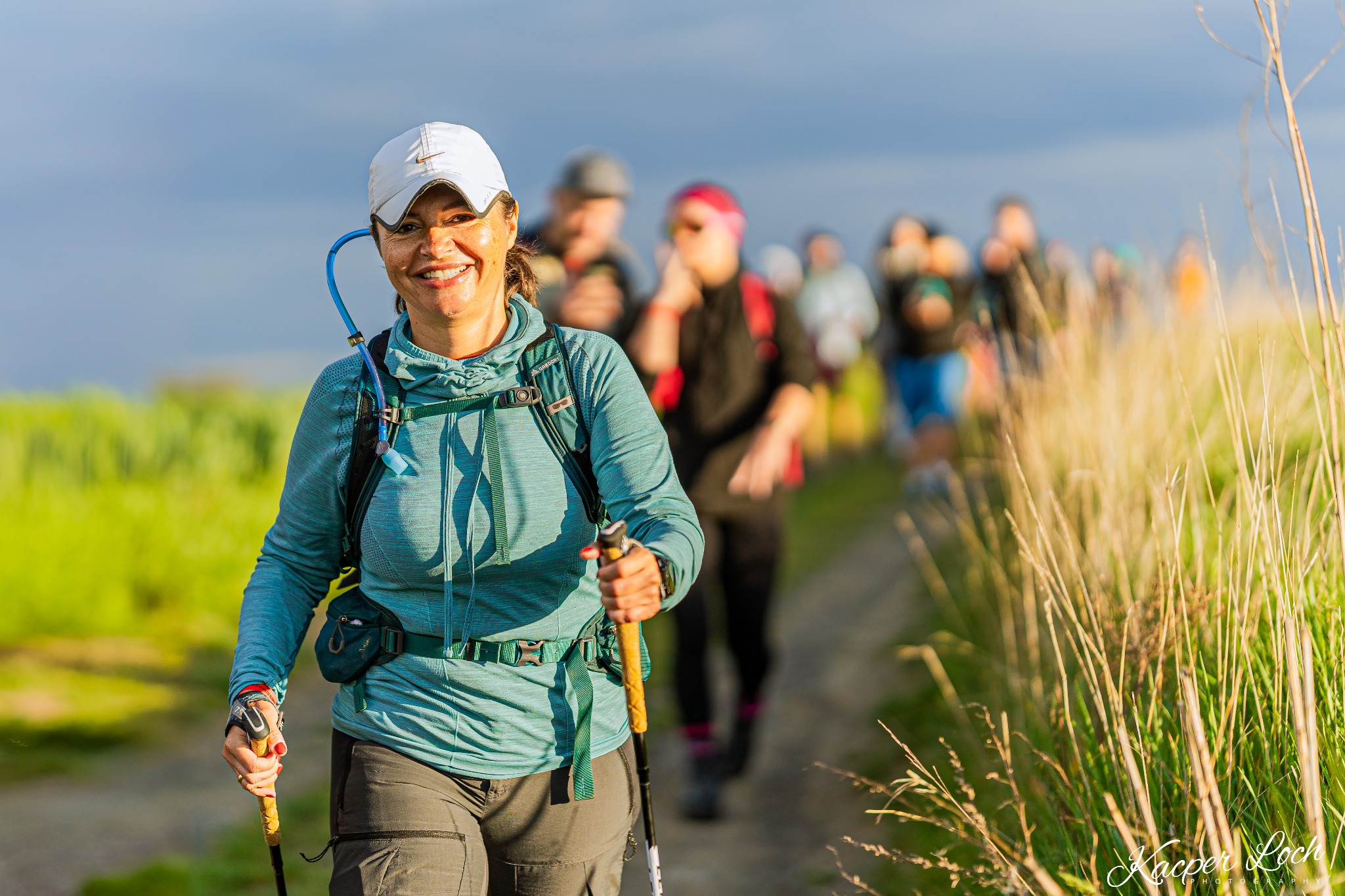 Uśmiechnięta kobieta z plecakiem i kijkami trekkingowymi prowadzi grupę osób polną ścieżką w słoneczny dzień.