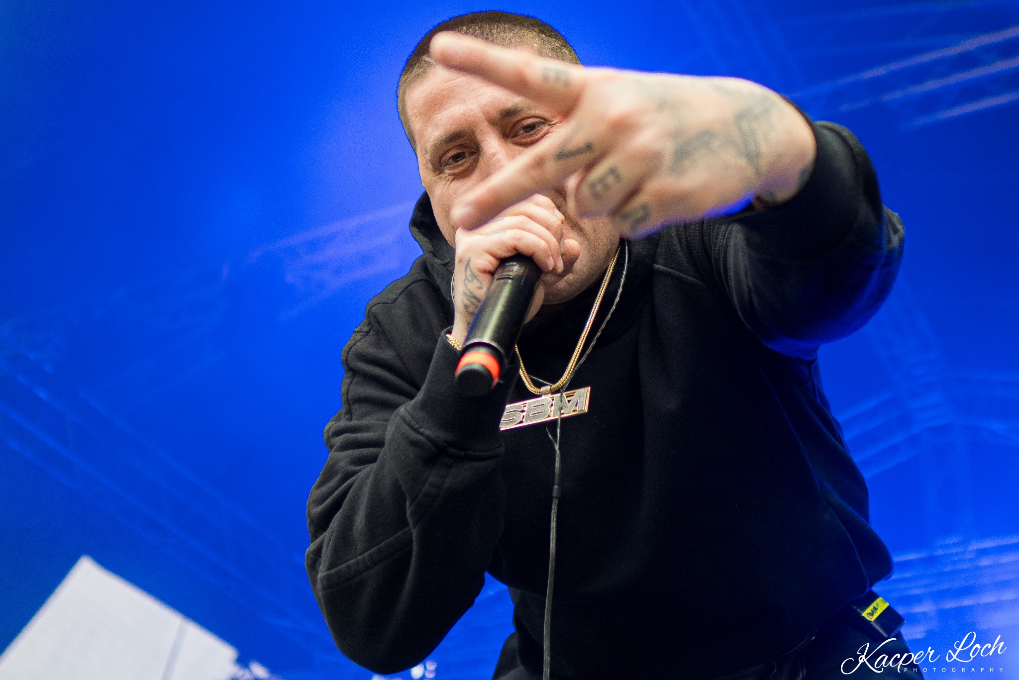 Tatuowany mężczyzna w czarnej bluzie z kapturem trzyma mikrofon i pokazuje znak V na tle niebieskiej ściany podczas występu.