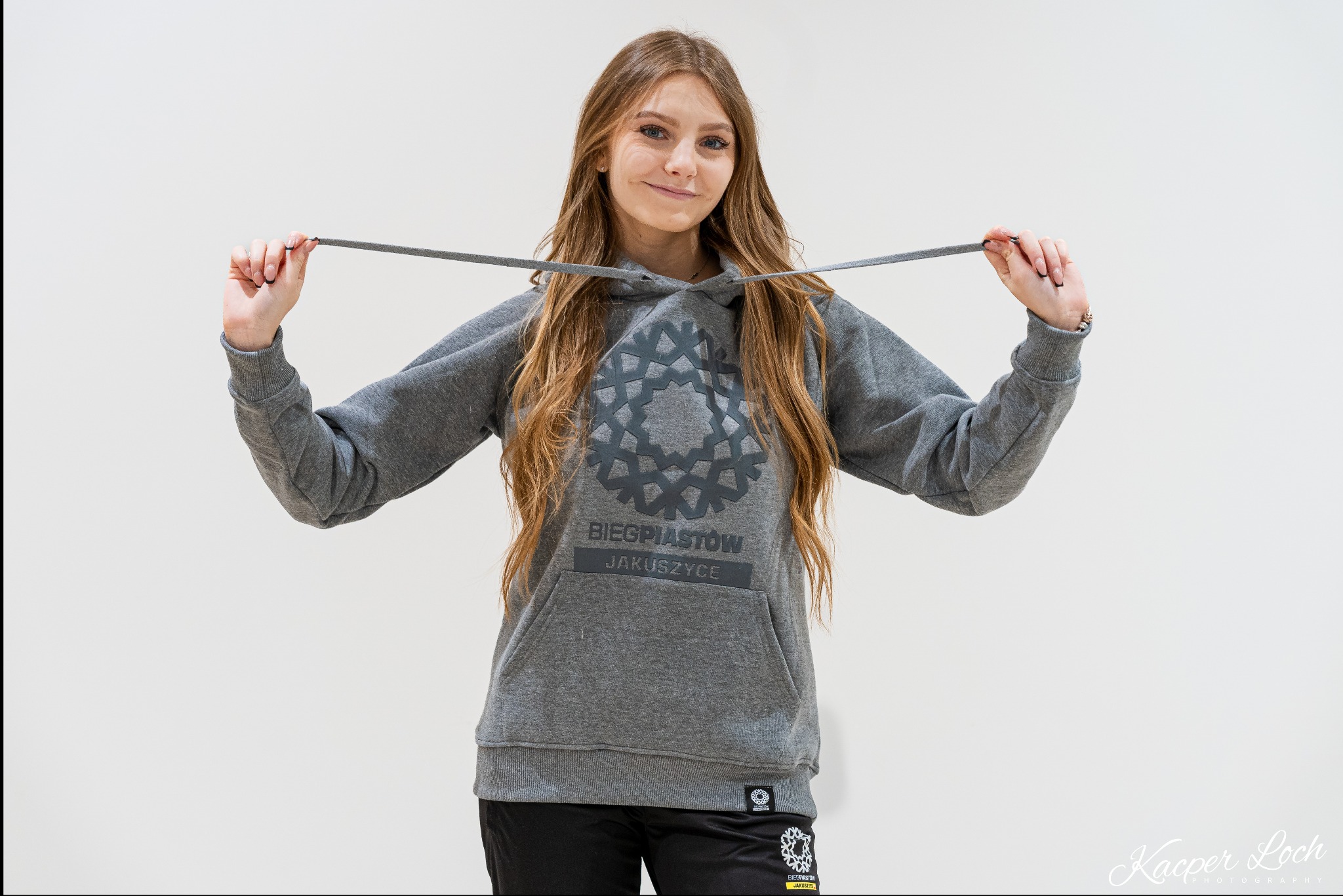 Młoda kobieta prezentuje szarą bluzę z kapturem z logo Biegu Piastów Jakuszyce na białym tle.