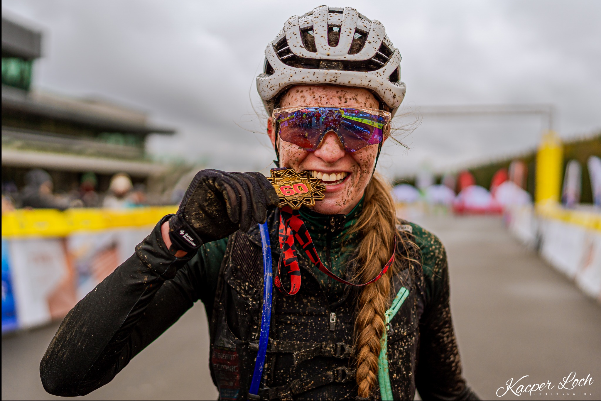 Uśmiechnięta, brudna od błota kobieta w kasku i okularach przeciwsłonecznych, trzymająca w dłoni medal z numerem 60 na tle mety wyścigu.