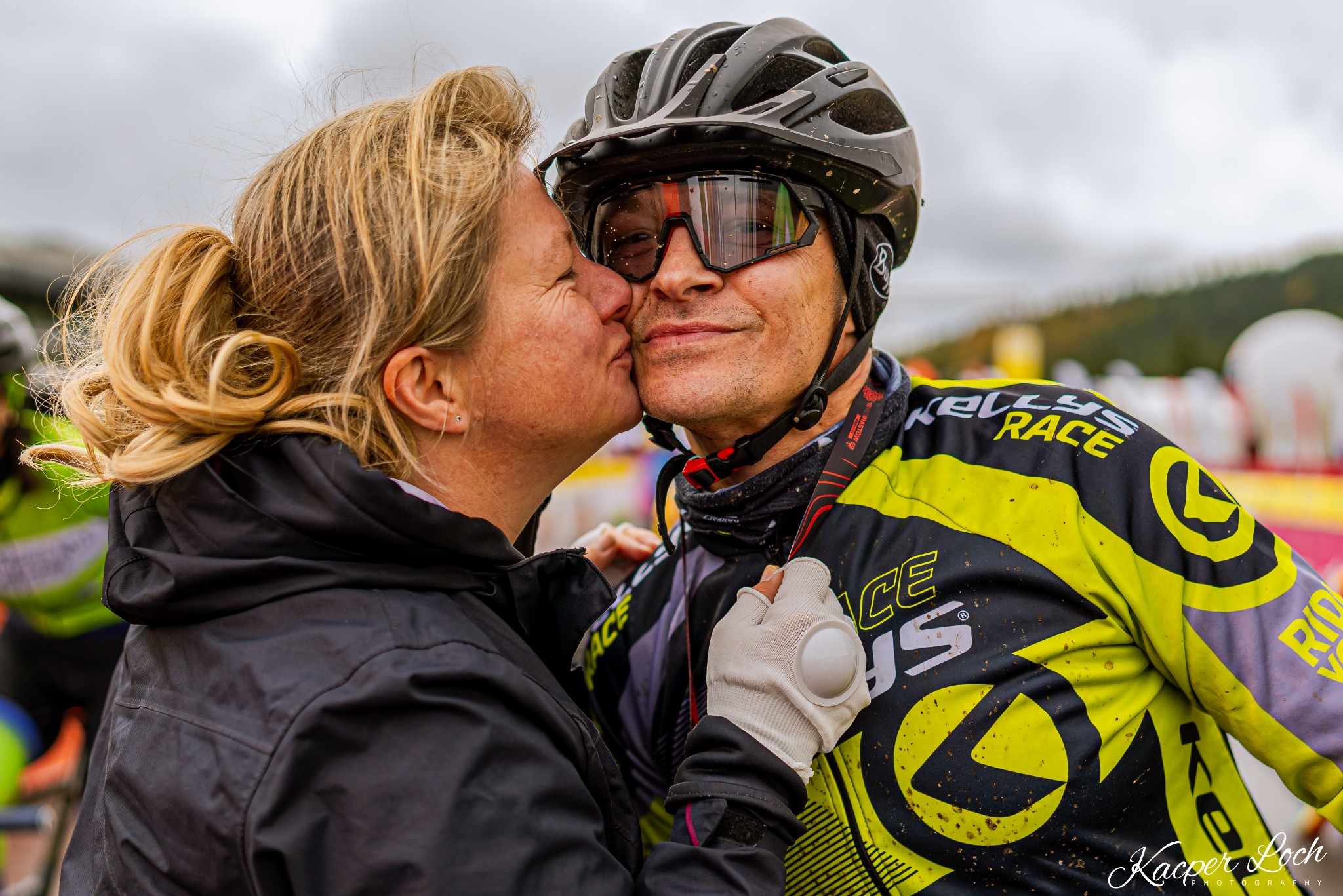 Kobieta całuje w policzek mężczyznę w kasku rowerowym i okularach przeciwsłonecznych, oboje ubrani w odzież sportową Kellys Race, widoczne zabrudzenia błotem.