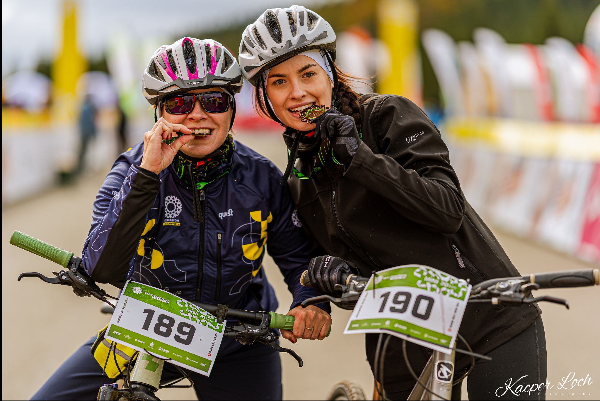 Dwie uśmiechnięte kobiety w kaskach rowerowych, trzymające w ustach i dłoniach medale po zawodach, z rowerami z numerami startowymi 189 i 190.