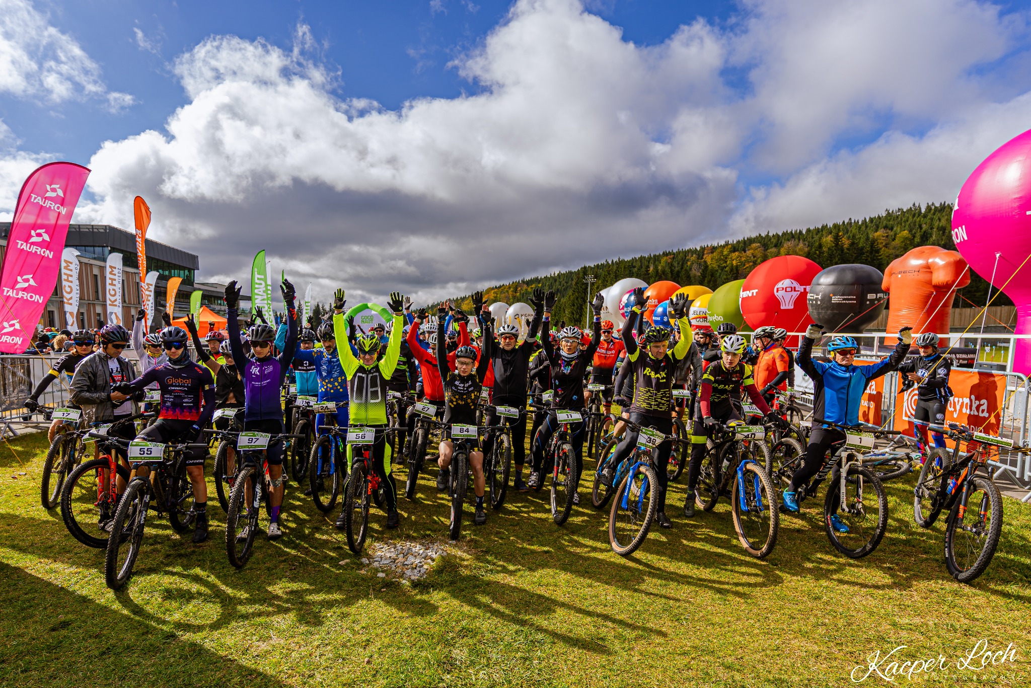 Grupa rowerzystów górskich z podniesionymi rękami na starcie wyścigu, widoczne flagi sponsorów i balony reklamowe.