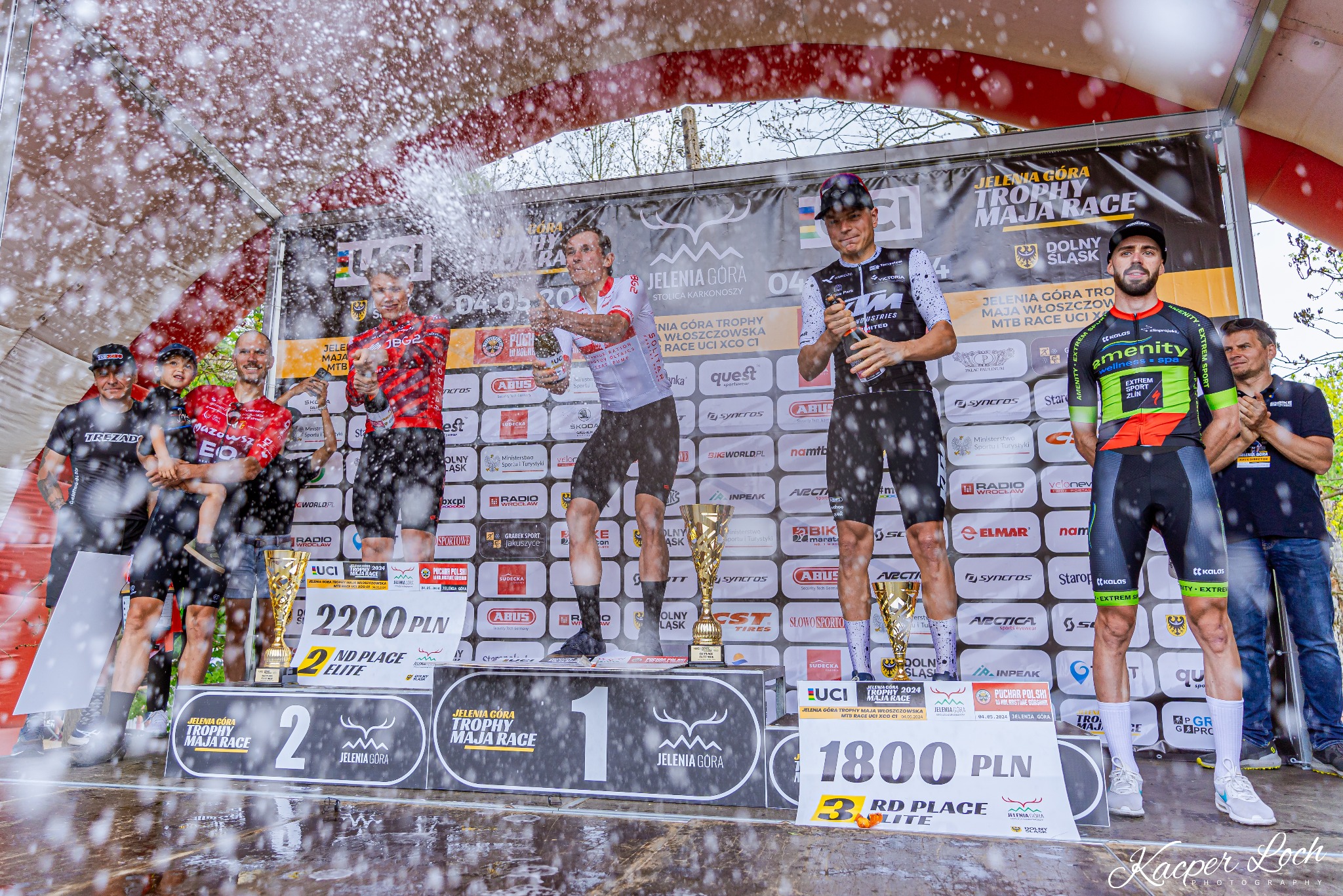 Zwycięzcy wyścigu rowerowego górskiego na podium, otwierają szampana, w tle banery sponsorskie i publiczność.