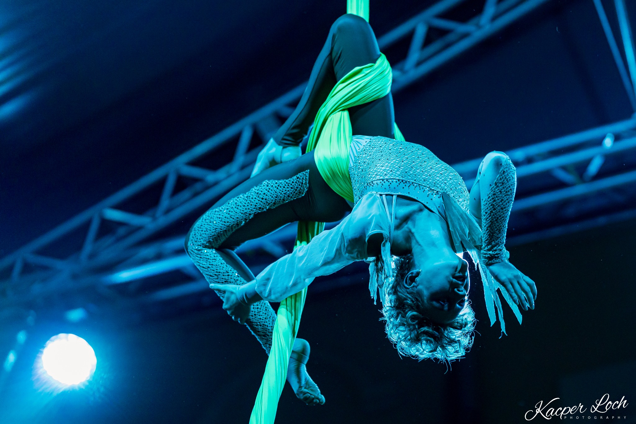 Akrobatka w kostiumie, zwisająca głową w dół na zielonej chuście, oświetlona niebieskim światłem reflektorów na tle konstrukcji scenicznej.