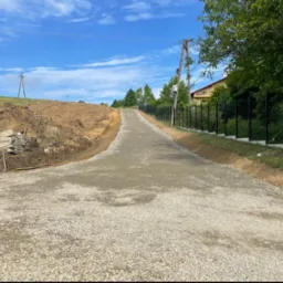 Droga gruntowa z kruszywa prowadząca pod górę, obok czarne ogrodzenie i wykopana ziemia, błękitne niebo z chmurami w tle.