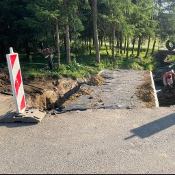 F.H.U KAM-DRÓG Kamil Mazur - Przygotowanie podłoża pod drogę: wykopany rów zabezpieczony czarną folią, robotnicy pracujący w wykopie, w tle mostek z oponami, znak ostrzegawczy po lewej stronie.